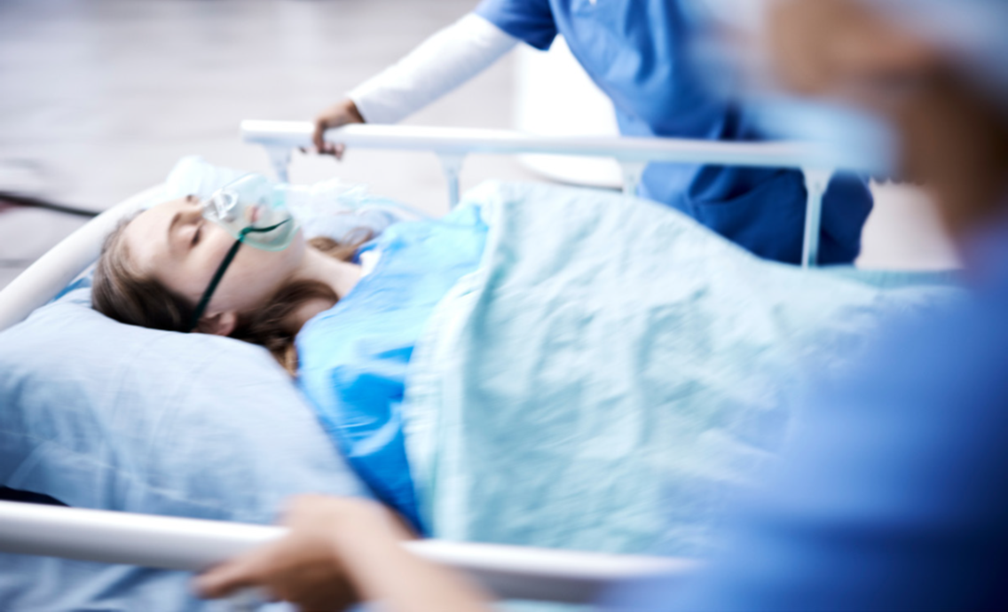 Bild: Ein M&auml;dchen liegt mit einer Atemmaske auf einem Krankenbett, das von zwei Personen in blauer Kleidung geschoben wird. Unsch&auml;rfe im Bild verdeutlicht die Schnelligkeit. von 