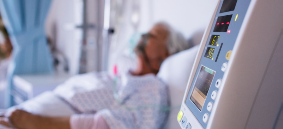 Bild: Frau mit Atemmaske in einem Krankenhausbett. Im Vordergrund steht ein Ger&auml;t zur &Uuml;berwachung der K&ouml;rperfunktionen.