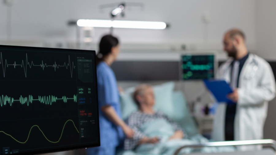 Bild: Ein Patient liegt im Krankenhausbett. Daneben stehen ein Arzt mit Klemmbrett und eine Schwester. Ringsum stehen Monitore, die K&ouml;rperfunktionen &uuml;berwachen..