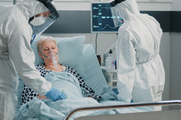 Bild: Eine &auml;ltere Frau liegt mit Beatmungsmaske in einem Krankenhausbett. Rechts und links davon stehen zwei Personen in Schutzkleidung. Eine untersucht die Frau, die andere schaut auf den &Uuml;berwachungsmonitor auf die K&ouml;rperfunktionen.