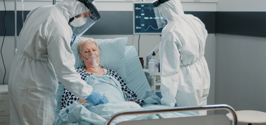 Bild: Eine &auml;ltere Frau liegt mit Beatmungsmaske in einem Krankenhausbett. Rechts und links davon stehen zwei Personen in Schutzkleidung. Eine untersucht die Frau, die andere schaut auf den &Uuml;berwachungsmonitor auf die K&ouml;rperfunktionen.